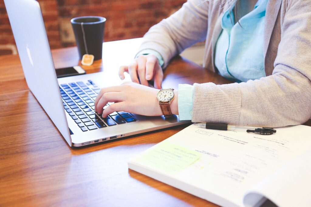 Foto de uma mulher com blusa bege e relógio marrom no braço, utilizando um computador cinza. Ao lado vemos um livro e uma xícara de café preta, em uma mesa de madeira (imagem ilustrativa). Texto: franquias virtuais 2021.