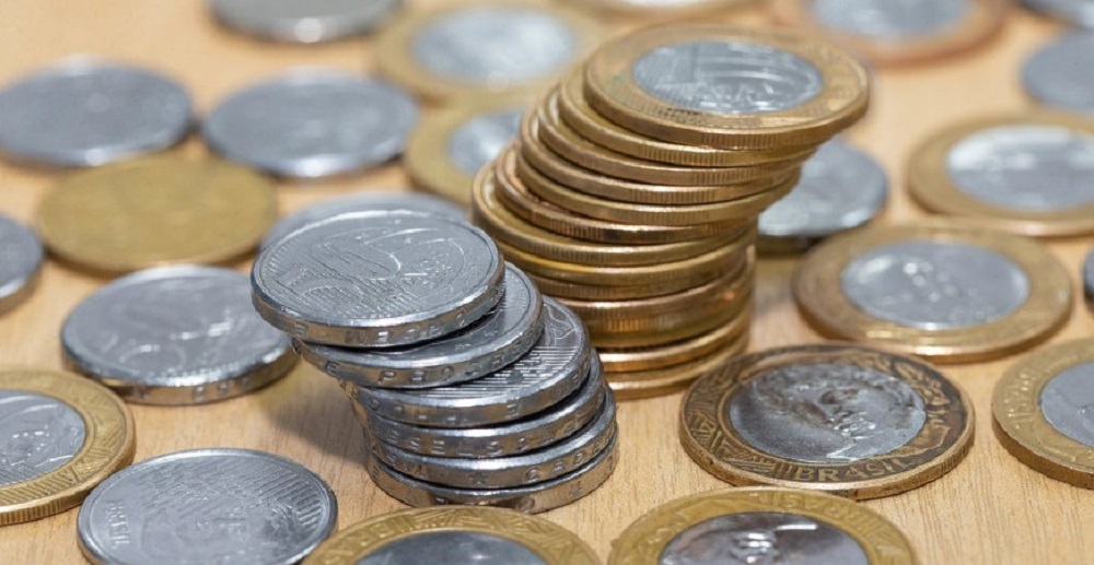 Pilha de moedas de um real e outra de moedas de cinquenta centavos e outras moedas iguais estas espalhadas ao redor. Imagem ilustrativa do texto abrir um negócio sem dinheiro.