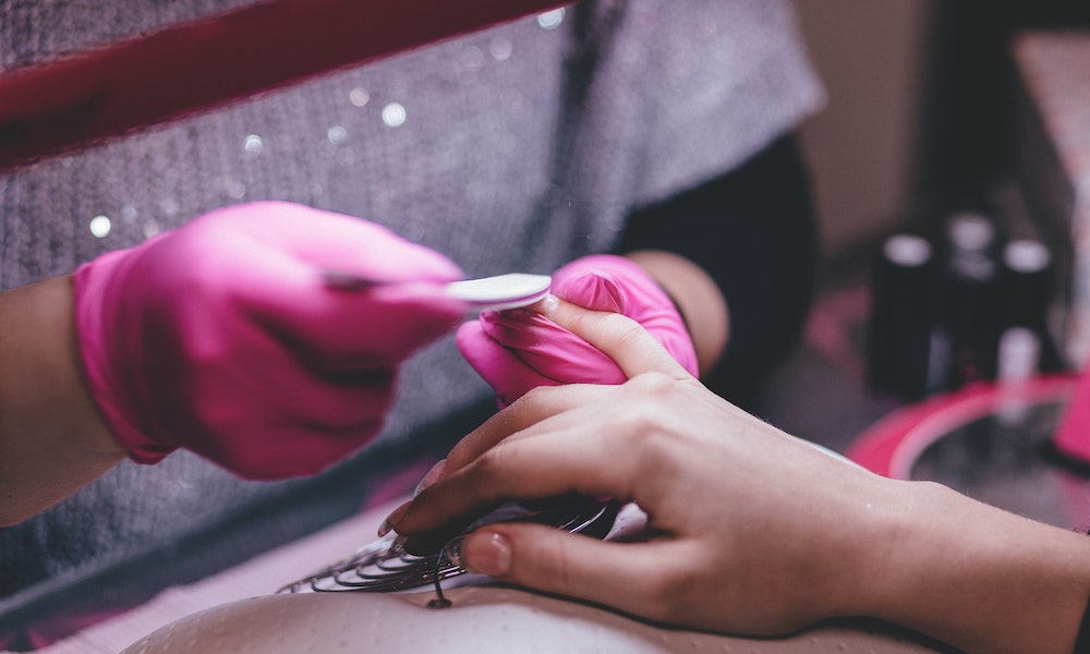Mãos com luvas de látex rosa lixam as unhas de uma mão feminina. Imagem ilustrativa entenda como funcionam as franquias de esmalteria.