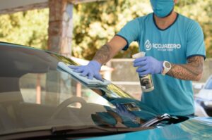 Homem usando máscara, e luvas na cor azul, limpando vidro de um carro. Ilustração do texto sobre franquia aplicativo delivery.