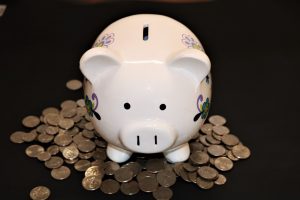 Imagem de um cofre em formato de porco na cor branca rodeado de moedas de dinheiro. Imagem ilustrativa texto como abrir uma franquia com pouco dinheiro.