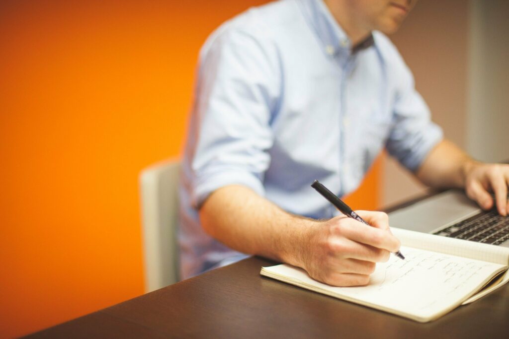 Foto de homem de blusa azul, utilizando um computador e escrevendo em um caderno. Ao fundo temos uma parede laranja. Imagem ilustrativa para texto franquias para trabalhar sozinho.