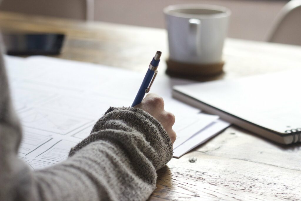 Mão de uma moça com blusa cinza, segurando uma caneta enquanto escreve em papéis. Ao fundo vemos uma caneca e um caderno em mesa de madeira. Imagem ilustrativa para texto franquias para trabalhar sozinho.