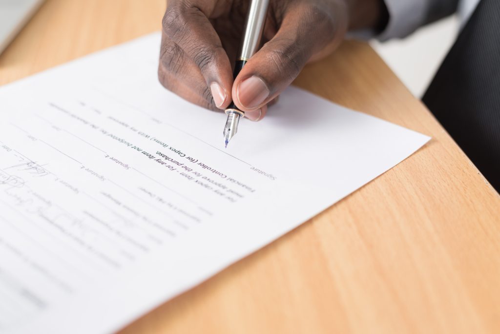 Imagem de da mão de um homem segurando uma caneta prestes a assinar um contrato (imagem ilustrativa). Texto: o que é royalties.