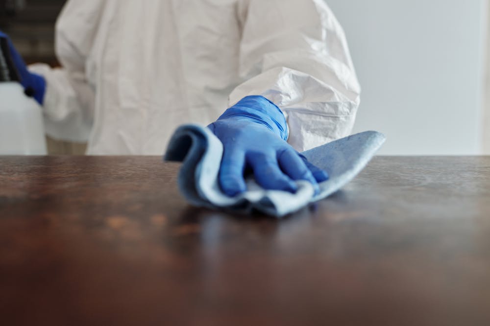 Pessoa usando luva azul, limpando superfície de mesa. Ilustração do texto sobre franquia de material de limpeza.