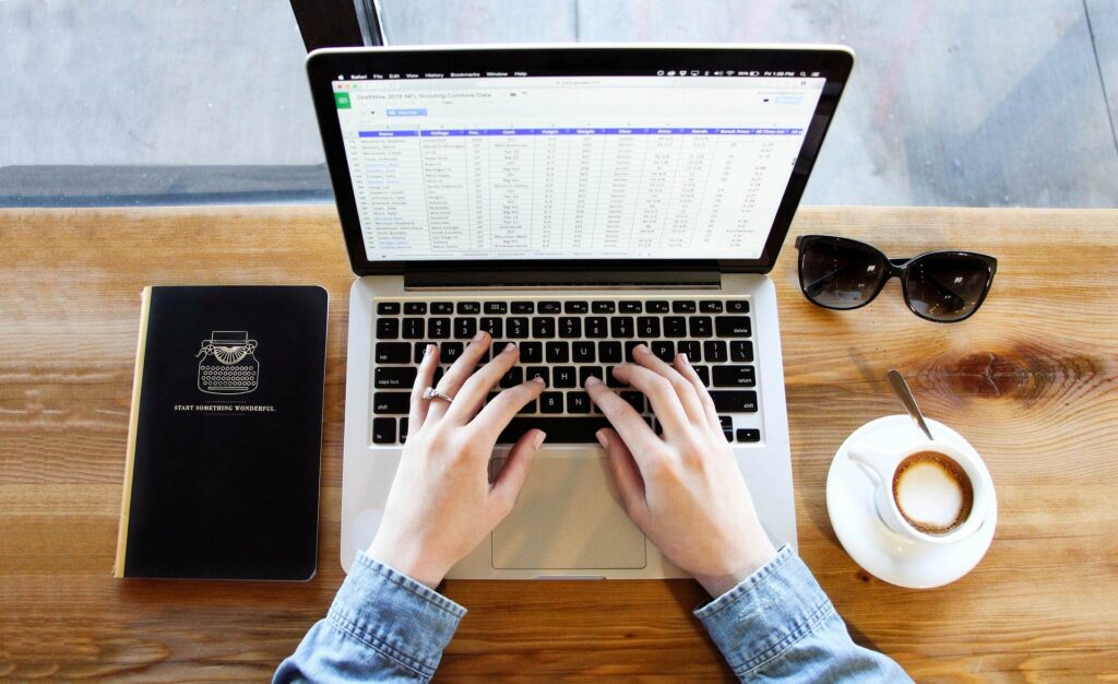 Pessoa de blusa jeans mexendo em um computador, em uma mesa de madeira. Temos um óculos escuros, xícara de café e caderno preto ao lado. Imagem ilustrativa para texto franquia de seguros barata.