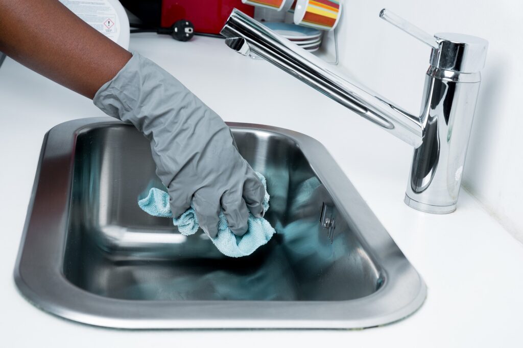 Foto da mão de uma pessoa com luva cinza enquanto limpa uma pia de metal com pano azul. Vemos um balcão branco e uma torneira de metal (imagem ilustrativa). Texto: franquias de limpeza e conservação.
