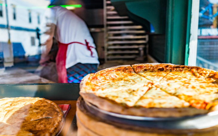 Imagem de uma pizza em destaque com pizzaiolo ao fundo quero abrir uma pizzaria como faço