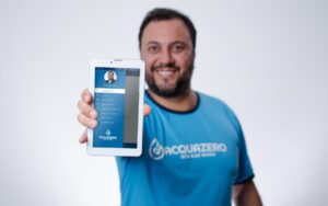 Imagem de um homem sorrindo, vestido com a camisa azul escrito Acquazero, segurando um tablet branco e mostrando a tela do para a câmera. Imagem ilustrativa do texto aplicativo Acquazero.