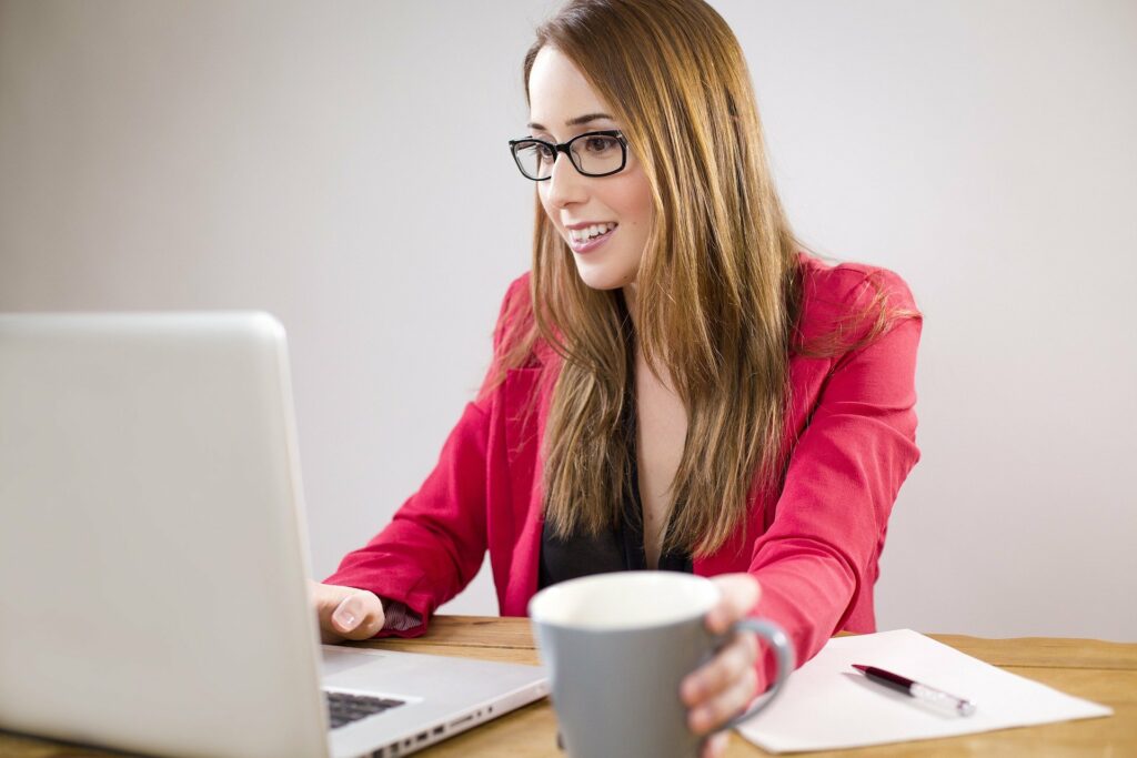 Vemos uma mulher de roupa rosa utilizando um computador, segurando uma xícara de café com um papel e caneta ao lado (imagem ilustrativa). Texto: franquias de seguros.