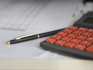 Foto de uma calculadora com botões vermelhos, em uma mesa branca, com uma caneta preta e alguns papeis. Imagem ilustrativa para texto franquia de contabilidade.
