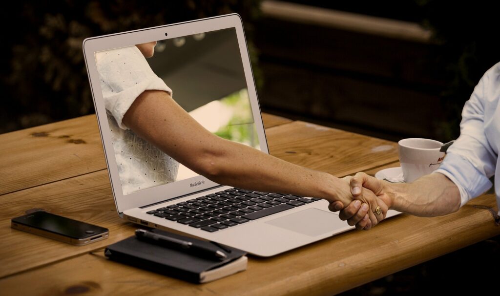Foto de um homem na frente do computador apertando a mão de um braço que está saindo de dentro do computador. Temos uma mesa de madeira, um celular e xícara branca. Imagem ilustrativa para texto abf franquias.