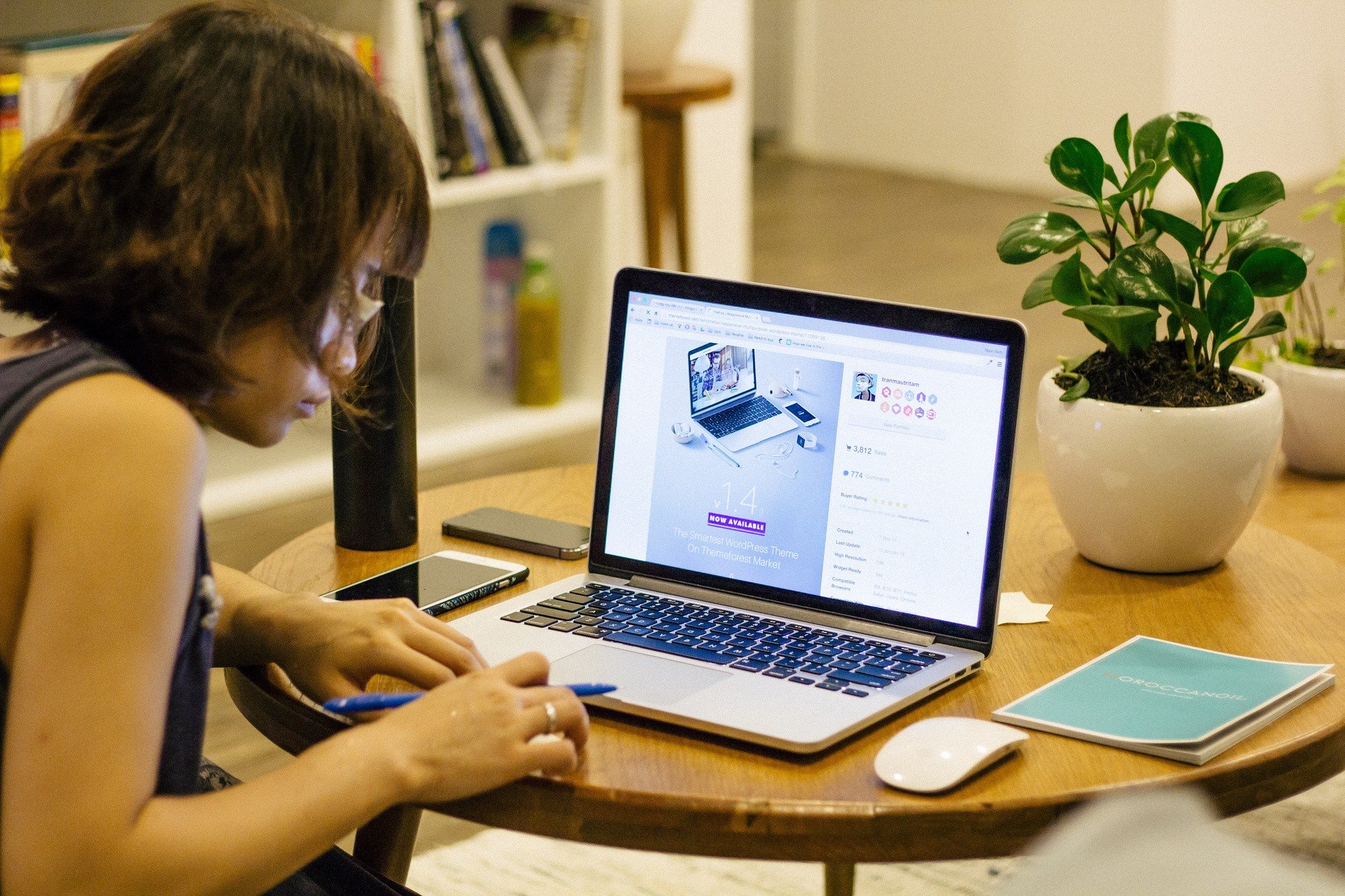 Foto de uma moça de blusa preta em uma mesa de madeira com um computador em cima. Ao lado vemos um caderno e um vaso de planta. Ao fundo vemos uma sala com uma estante branca. Imagem ilustrativa para texto franquias 2021 tendências.