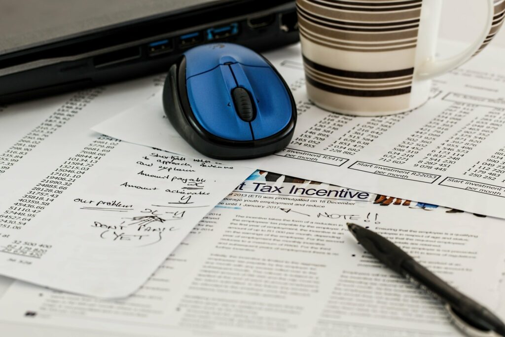 Foto de vários papeis em uma mesa, com parte de um computador, um mouse, uma caneca e uma caneta (imagem ilustrativa). Texto: franquia de contabilidade.