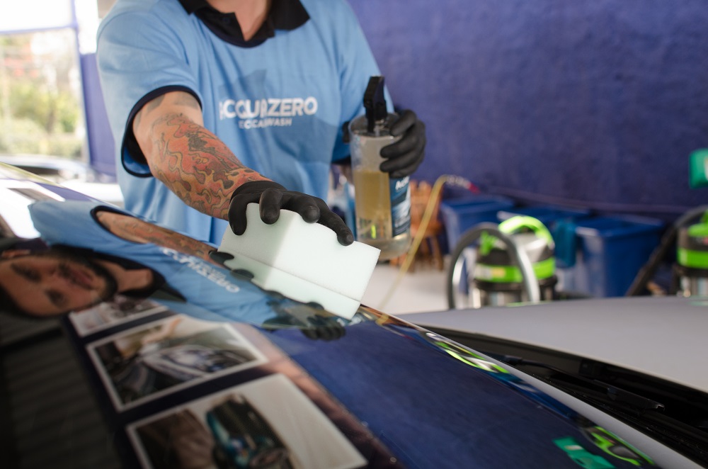 Funcionário da Acquazero com o uniforme da empresa fazendo a limpeza do vidro de um carro. Ao fundo vemos uma parede azul e equipamentos. Imagem ilustrativa para o texto franquia de estética automotiva.