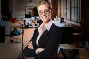 Mulher empreendedora de óculos com um escritório ao fundo.