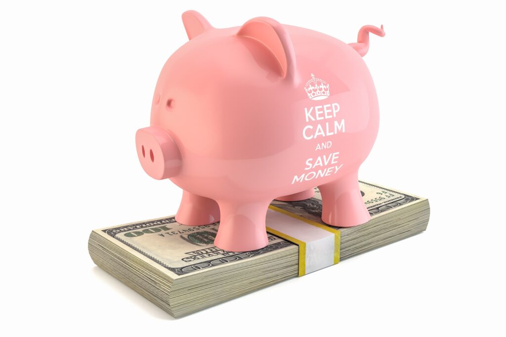 Vemos um cofrinho  rosa em forma de porco em cima de um maço de notas de dólar (imagem ilustrativa). Texto: como empreender com pouco dinheiro.
