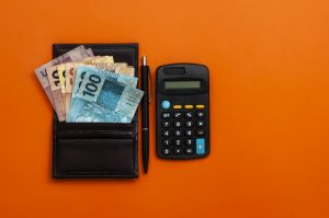 Imagem de uma carteira com várias notas de dinheiro ao lado de uma calculadora preta e um fundo laranja na imagem. Imagem ilustrativa texto abrir uma financeira.