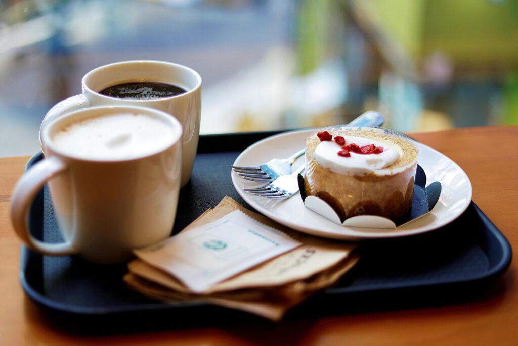 Foto de uma bandeja com dois café e um bolo ao lado, em uma mesa marrom (imagem ilustrativa). Texto: franquia de bebidas.