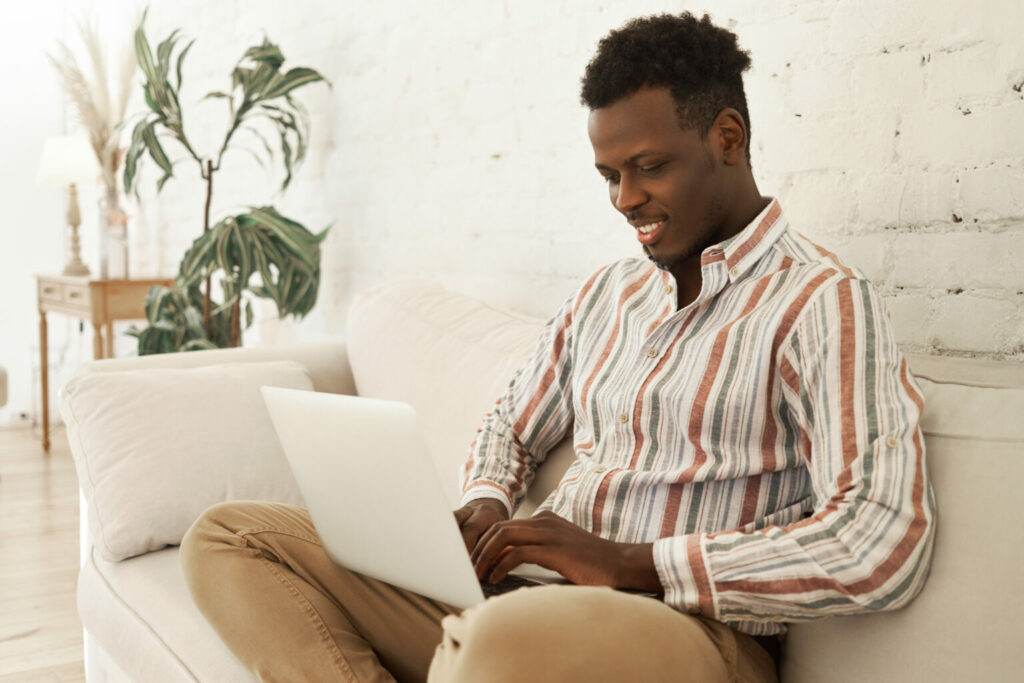 Vemos um homem negro sentado no sofá de casa enquanto usa seu laptop.
