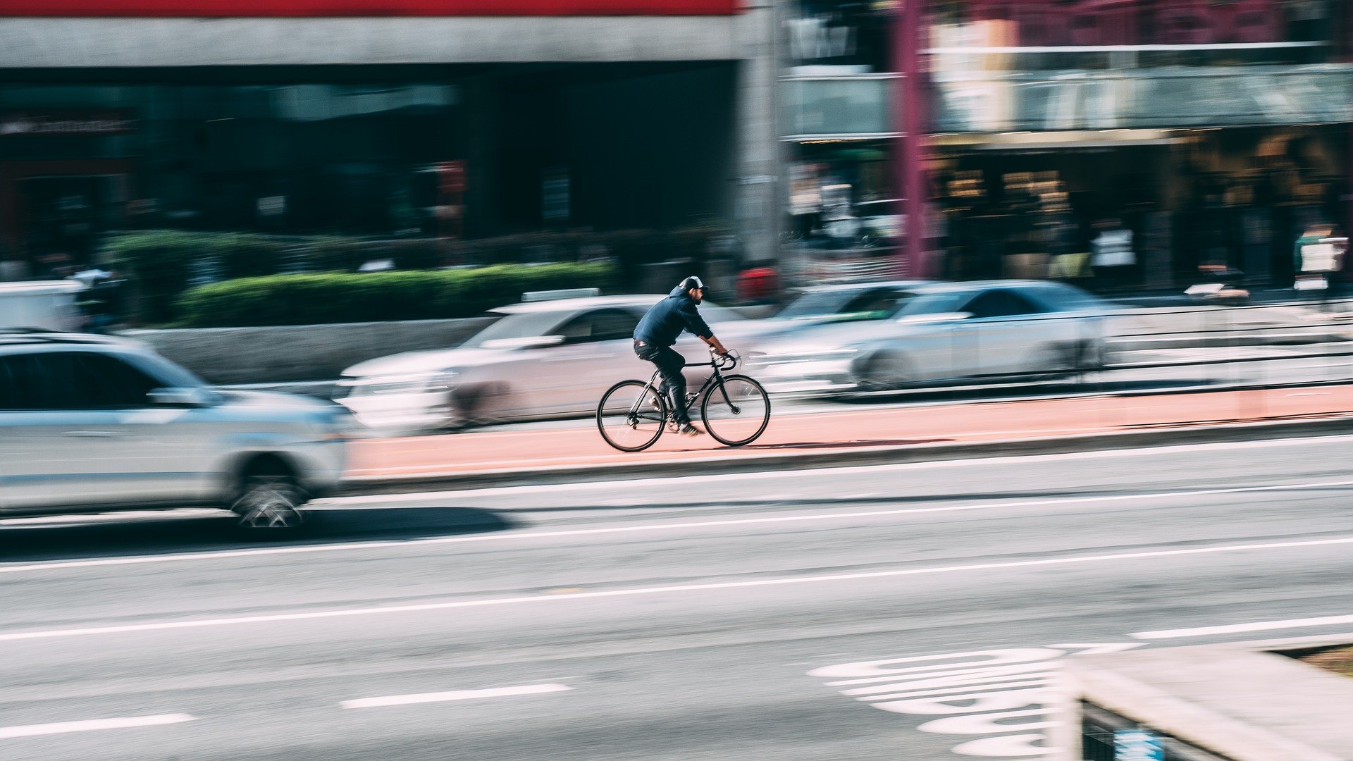 Vemos um ciclista no meio de uma avenida com lojas e carros ao redor (imagem ilustrativa). Texto: ideias de negócios com pouco dinheiro.