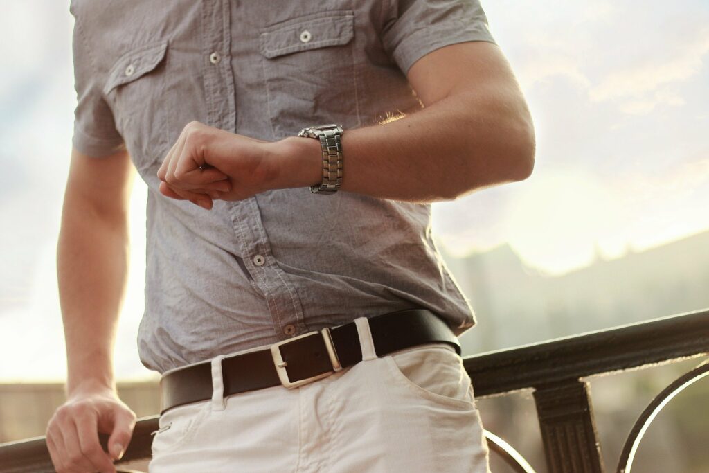 Homem de blusa cinza e calça branca, olhando as horas em um relógio de braço. 