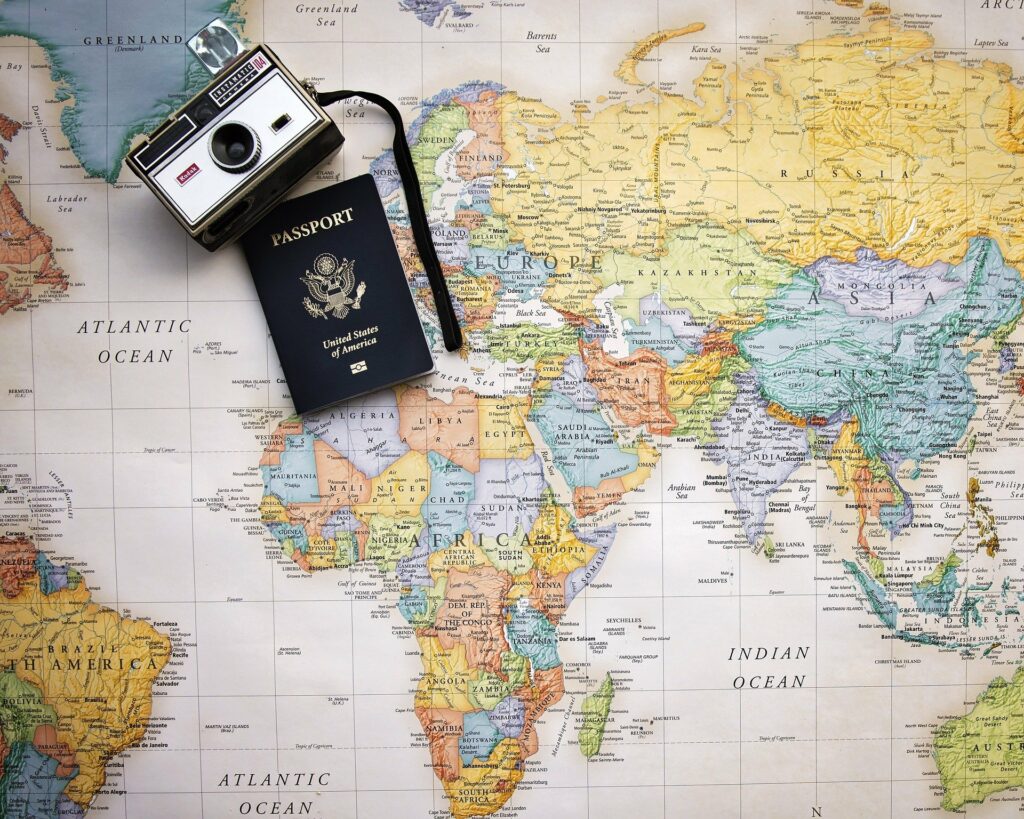 Vemos a imagem de de um mapa-múndi ao fundo com um passaporte e uma câmera em cima. 
