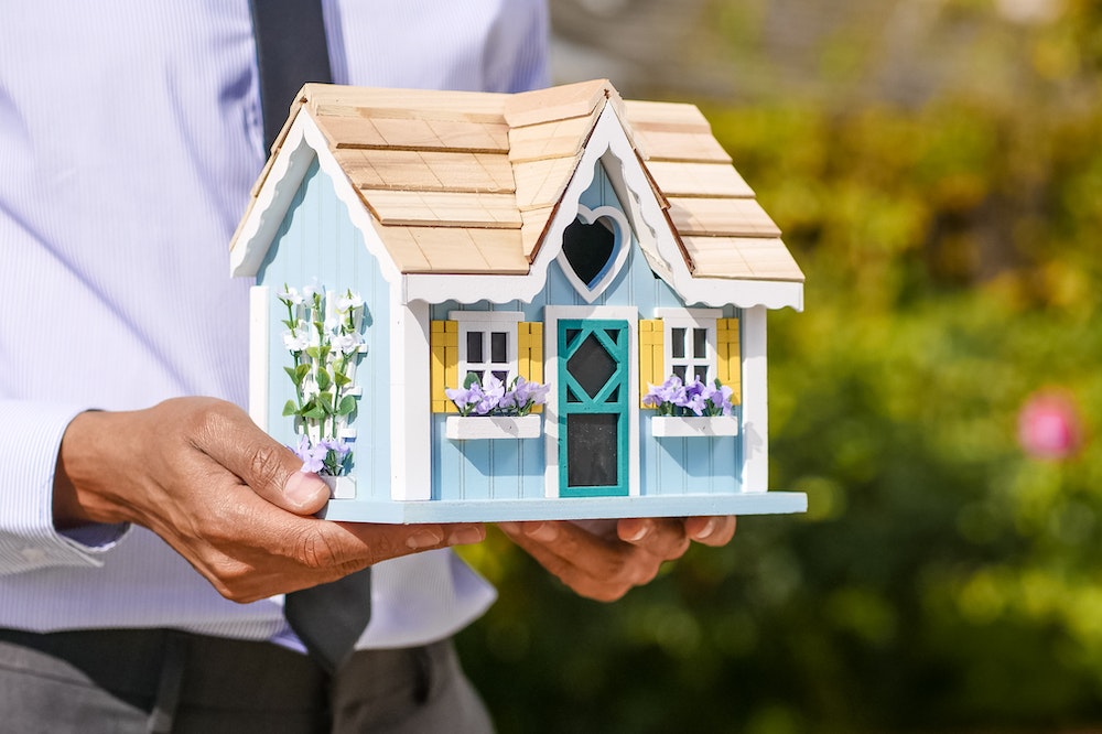 Homem usando camisa social azul, segurando uma casinha de brinquedo. Ilustração do texto sobre franquia Quisto se destaca em setor de seguro residencial.