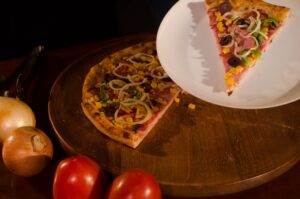 Fatia de pizza a moda em um prato branco e uma outra parte da pizza em uma tábua de madeira com tomates e cebolas ao redor. Imagem ilustrativa do texto opções de franquias de pizzaria.
