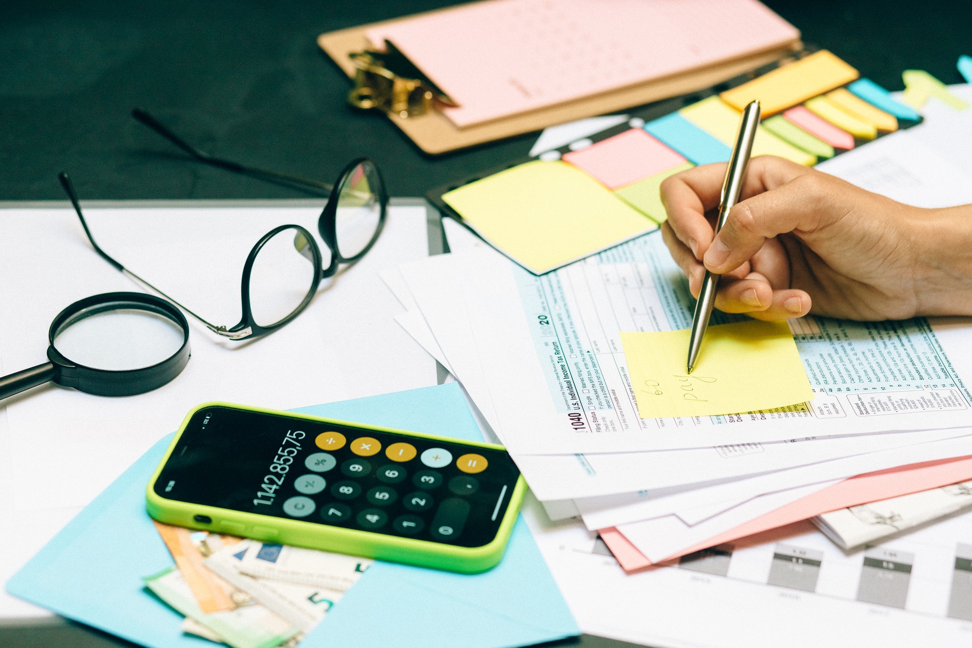 Imagem de uma mesa cheia de papeis, uma calculadora, um óculos e uma mão escrevendo em um papel. Imagem ilustrativa texto afinal o que é franquia.