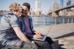 Homens sorridentes, sentados lado a lado ao ar livre, trabalhando em um notebook cinza. Ilustração do texto sobre empreender ou investir.