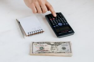 Imagem de uma mão usando uma calculadora e um bloco de notas e notas de dólar ao lado. Imagem ilustrativa texto negócios com baixo investimento.