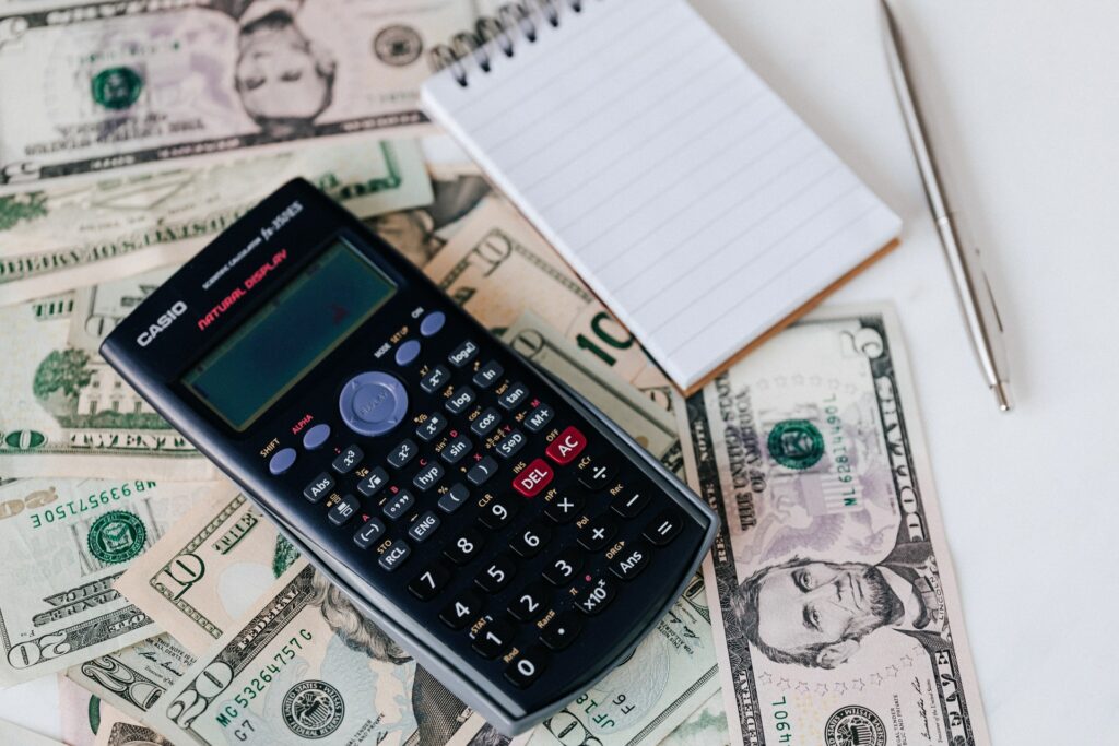 Imagem de uma calculadora em cima de notas de dólares e um caderno de anotação ao lado. Imagem ilustrativa texto expandir seu negócio.