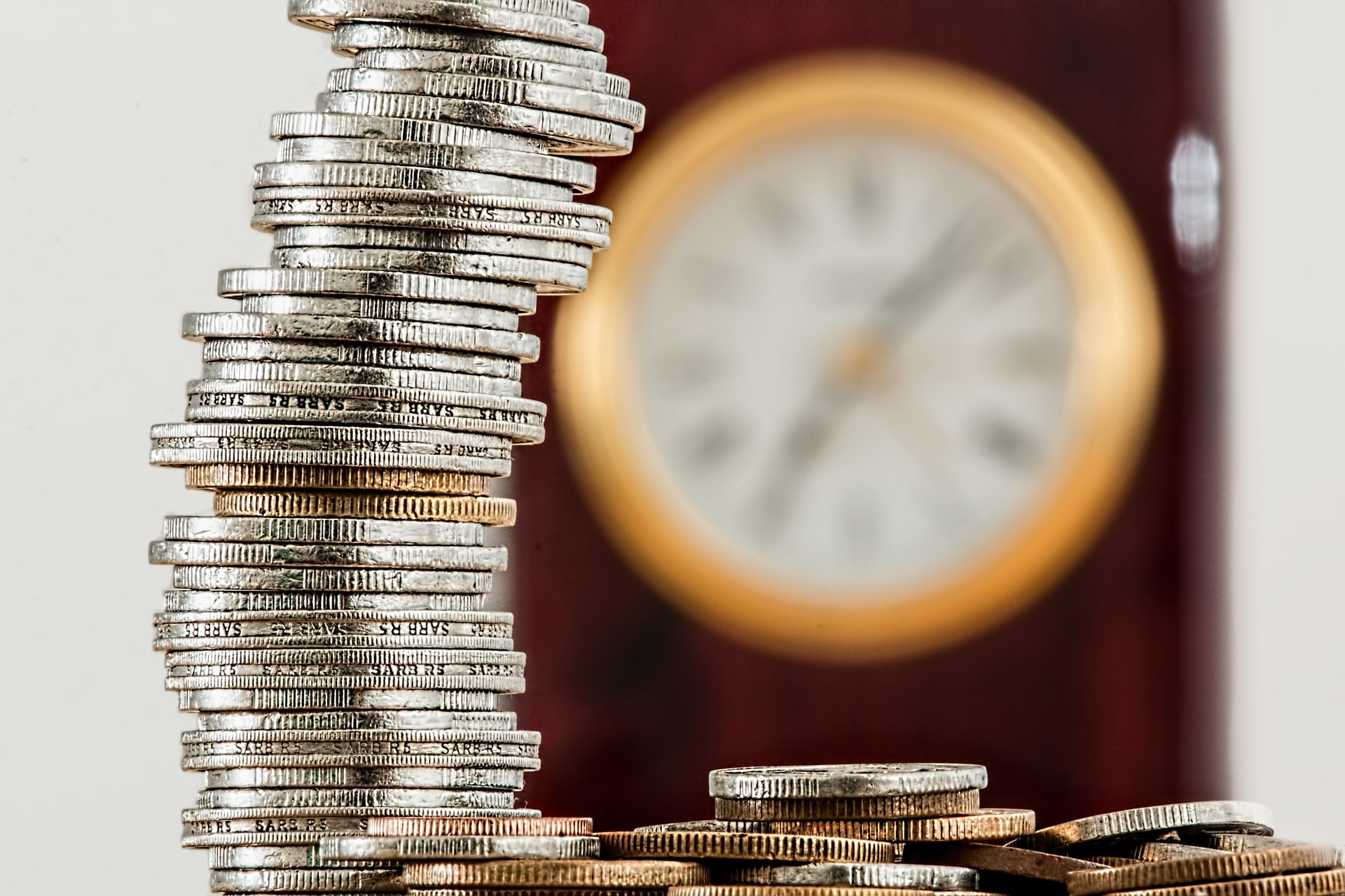 Imagem de uma pilha de moedas com um relógio ao fundo. Imagem ilustrativa texto investimentos a longo prazo.