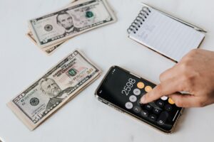 Imagem de uma mão mexendo na calculadora do celular em uma mesa com notas de dinheiros e um caderno de anotações. Imagem ilustrativa texto melhores setores para investir.