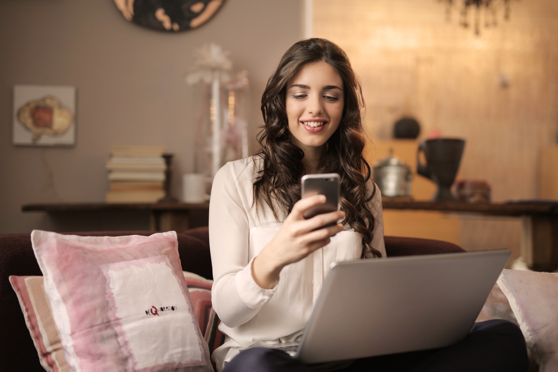 Imagem de uma mulher sentada no sofá com um computador no colo e um celular na mão. Imagem ilustrativa texto franquias online baratas e lucrativas.