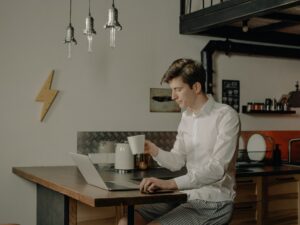Imagem de um homem segurando uma xícara na mesa da cozinha em frente um computador. Imagem ilustrativa texto microfranquia home office.