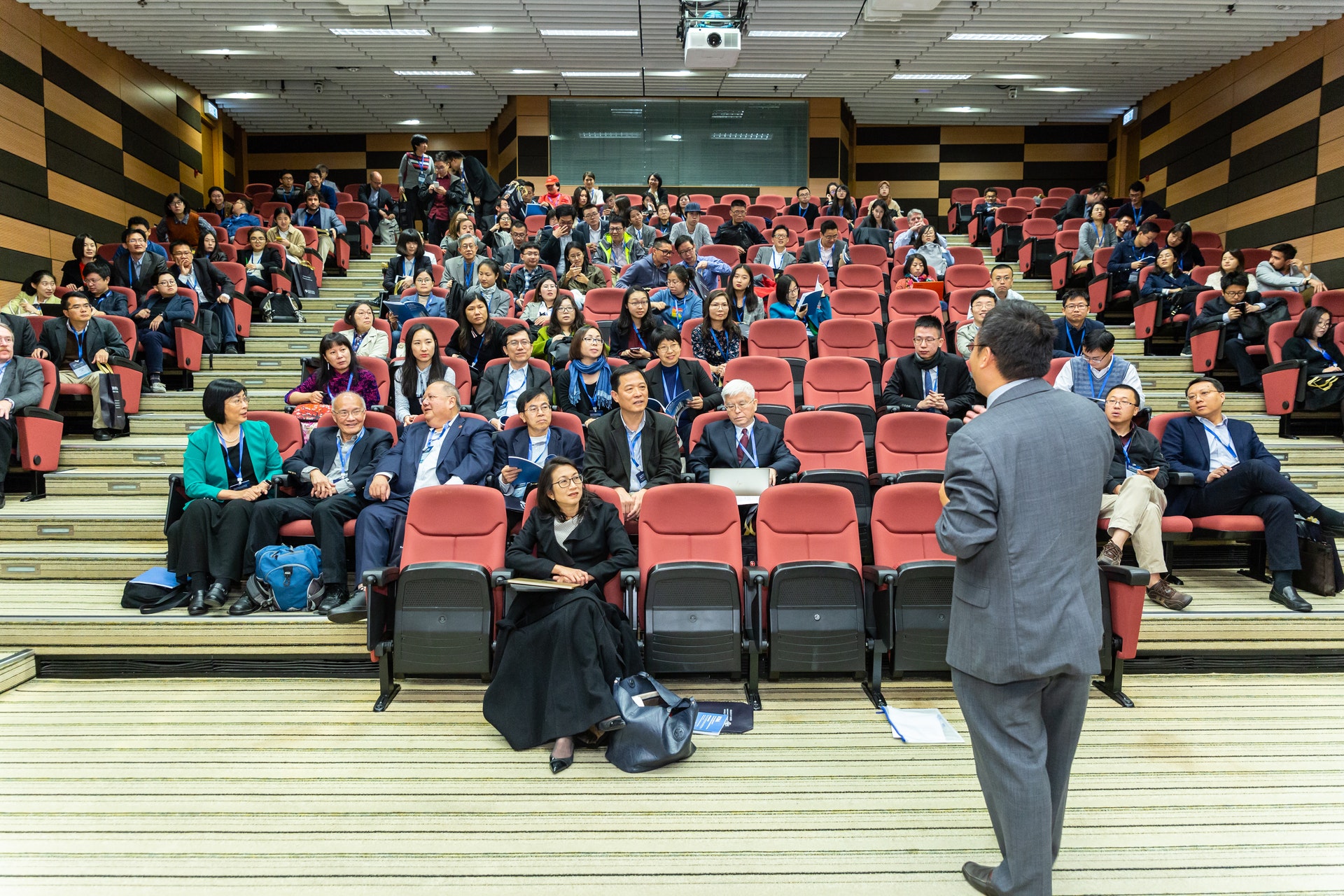 Imagem de um salão de palestra cheio de pessoas nas cadeiras e uma pessoa falando no palco. 