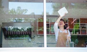 Mulher de camisa branca limpa as vidraças da janela. Imagem ilustrativa texto franquias de limpeza e conservacao.