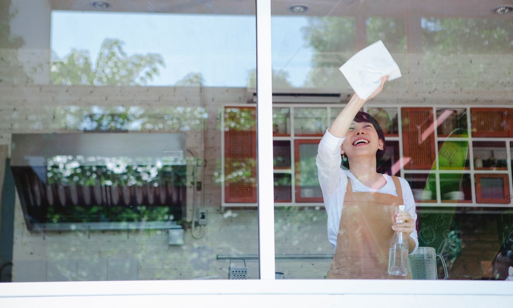 Mulher de camisa branca limpa as vidraças da janela. Imagem ilustrativa texto franquias de limpeza e conservacao.