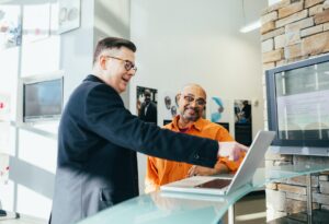 Imagem de um empreendedor sorrindo apontando para um computador junto a outro homem. Imagem ilustrativa do texto grandes empreendimentos.