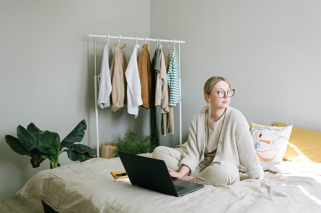 mulher usando notebook sentada em cama em quarto com cabideiro ilustração de empreendedorismo digital