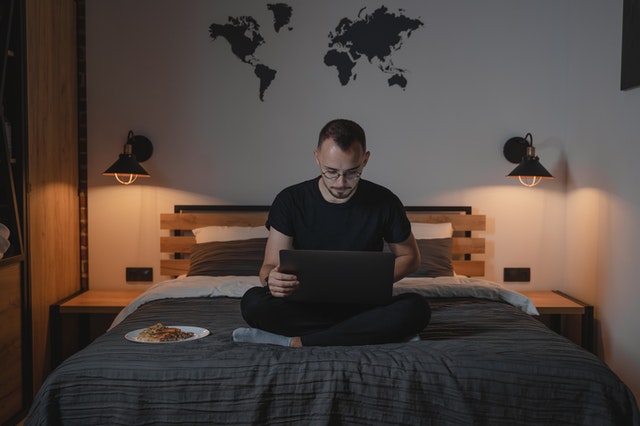 homem usando notebook que está em seu colo sentado na cama, com mapa mundi atrás