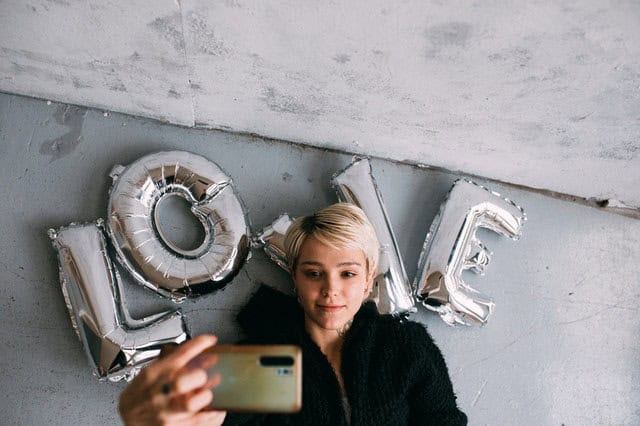 Mulher deitada com balões em formato de letras, formando a palavra love atrás e tirando selfie com celular.