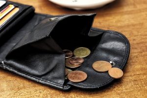 Imagem de uma carteira preta aberta sobre uma superfície de madeira com várias moedas saindo dela. Imagem ilustrativa texto como abrir um negócio sem dinheiro.