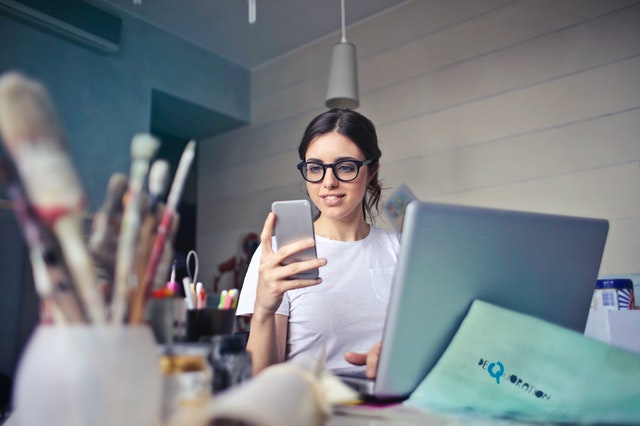 Mulher usando um celular em frente a notebook e pinceis em escritório. Ilustração do texto sobre ideias de negócios para mulheres.