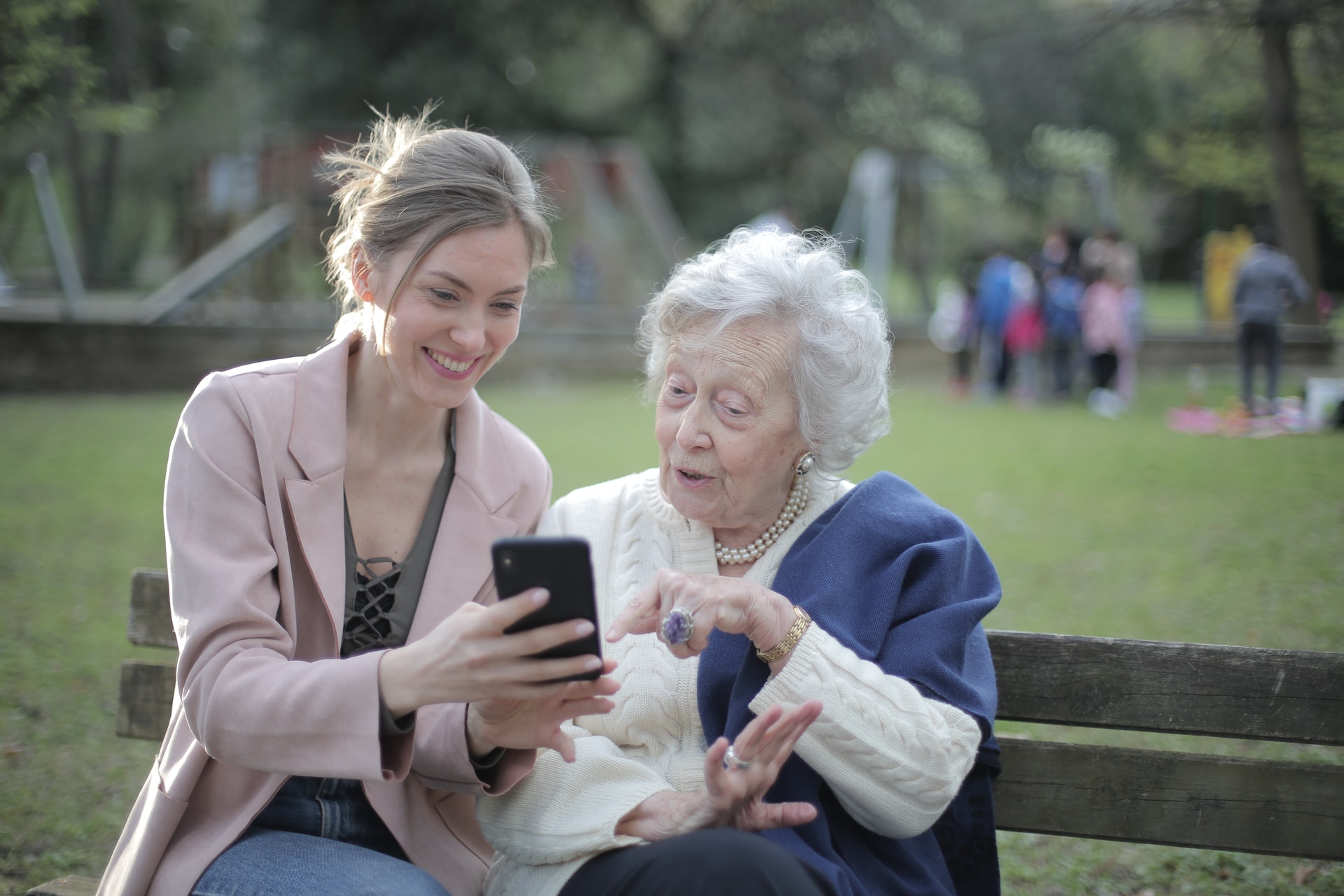 Mulher e uma idosa olhando para um celular sentadas em um banco de parque. 