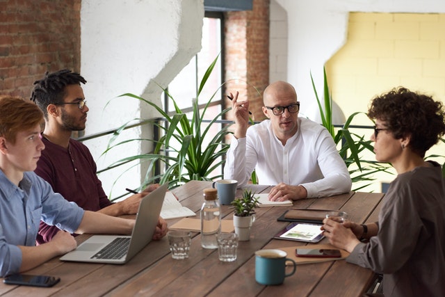 Reunião de negócios com grupo de quatro pessoas em mesa de madeira com cafés e computadores.