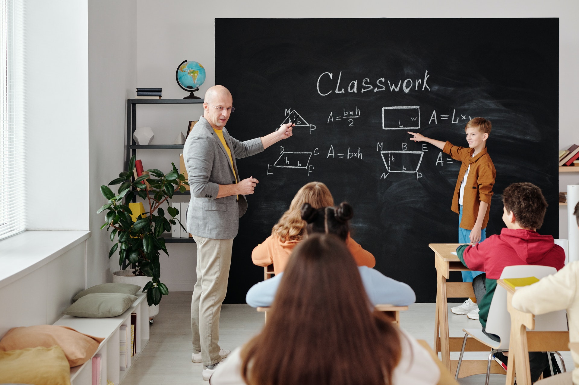 Professor em frente aos alunos em uma sala mostrando algo em um quadro e ao lado de uma criança em pé. Imagem ilustrativa do texto curso de inglês para crianças.