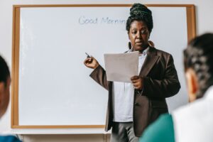 Professora segurando um papel em frente a um quadro branco escrito Good Morning. Imagem ilustrativa do texto curso de inglês para crianças.